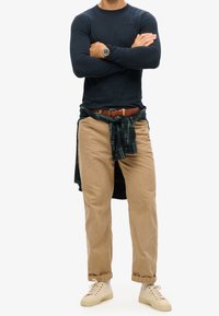 Marineblaues Langarmshirt kombiniert mit beigen Cordhosen. Karohemd um die Taille gebunden, brauer Gürtel und beige Sneaker. Klassischer Schnitt.