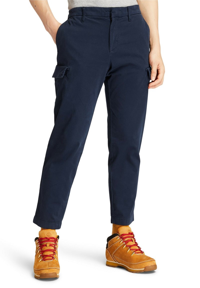 Personne portant un pantalon cargo bleu marine et des chaussures de randonnée jaune moutarde avec des lacets rouges, debout les mains dans les poches.