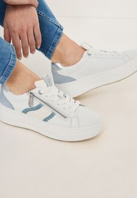 Sneakers bianchi con dettagli in denim, tacco argento e zip laterale. Presentano punta rotonda, suola piatta e design con lacci. Jeans blu visibili.