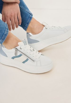 White sneakers with denim accents, a silver heel, and a side zipper. Features round toe, flat sole, and lace-up design. Blue jeans visible.
