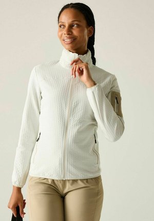 Mujer sonriente, vestida con una chaqueta blanca texturizada con cremallera y pantalones beige, de pie frente a un fondo claro y liso.