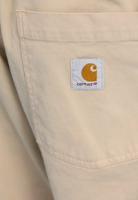 Beige fabric with a smooth texture, featuring a Carhartt logo patch in orange and white at the back pocket.