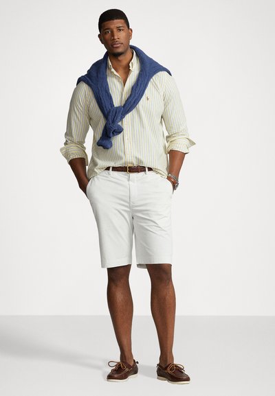 Λευκά σορτς, ανοιχτό κίτρινο πουκάμισο με ρίγες, ναυτικό πουλόβερ δεμένο στους ώμους, καφέ ζώνη, καφέ παπούτσια με κορδόνια, σε στάση όρθιος.