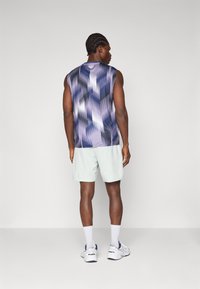 Ärmelloses Sportoberteil im lila und schwarzen geometrischen Muster, kombiniert mit hellgrünen Shorts. Model trägt weiße Sneakers und Socken.