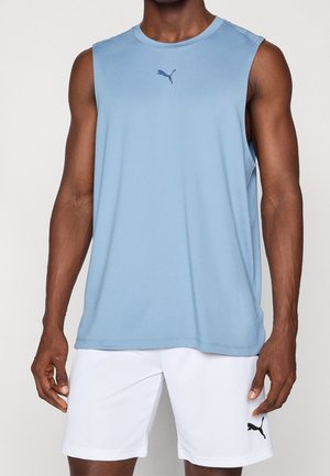 Man draagt een lichtblauw mouwloos sportshirt en witte short met Puma-logo's, staand tegen een effen lichte achtergrond.