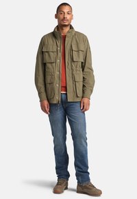 Chaqueta verde oliva con cierre frontal, cuatro bolsillos en la parte delantera y un cuello alto, combinada con una camisa color óxido y jeans azules.