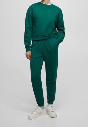 Personne portant un sweat-shirt vert foncé et un pantalon de jogging assorti avec des chaussettes blanches et des baskets blanches, debout devant un fond uni.