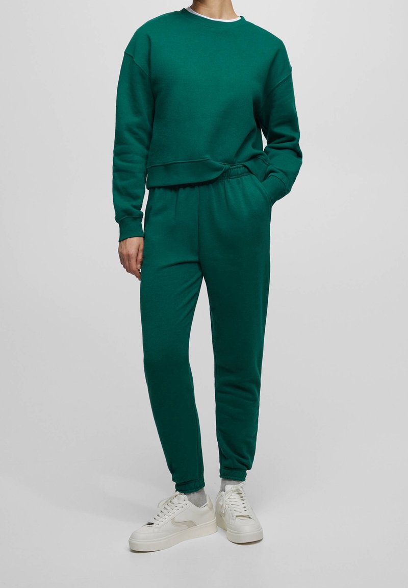 Personne portant un sweat-shirt vert foncé et un pantalon de jogging assorti avec des chaussettes blanches et des baskets blanches, debout devant un fond uni.
