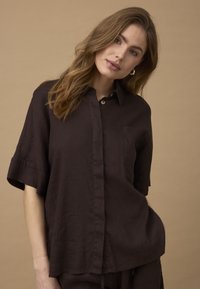 Vrouw met golvend lichtbruin haar, gekleed in een losse donkerbruine blouse met korte mouwen en knopen, staand tegen een beige achtergrond.
