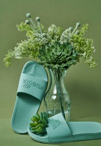 Para miętowych klapek wsuwanych o teksturowanej wkładce i logo marki, obok szklanej wazy z zielonymi liśćmi i kwiatami.