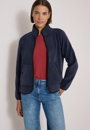 Femme portant une veste zippée bleu marine par-dessus un t-shirt rouge et un jean bleu clair, debout devant un fond clair uni.