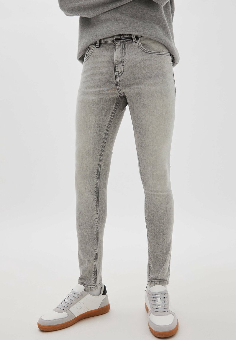 Personne portant un jean skinny gris clair et des baskets blanches avec des semelles marron, debout devant un fond clair uni.