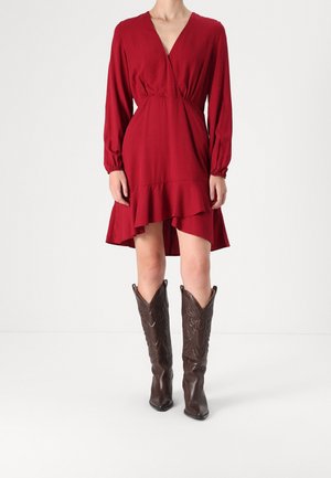 Femme portant une robe portefeuille rouge longueur genoux avec un ourlet à volants et des manches longues, associée à des bottes de cowboy marron foncé à motifs.