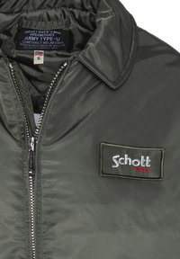 Roheline nailonist pommitaja jakk, millel on lukuga sulgemine, krae ja ristkülikuline Schotti logo patch. Sile tekstuur, klassikaline sõjaväe disain.