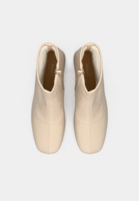 O pereche de cizme bej până la glezne cu fermoare pe laturile interioare, căptușeală moale și vârfuri pătrate, văzute din sus.