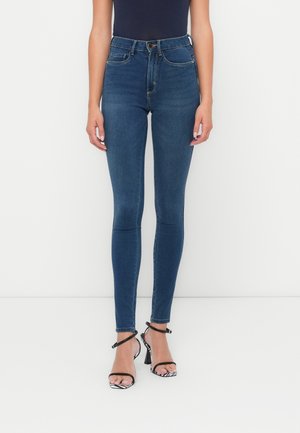 Jean bleu taille haute, ajusté, avec un design classique à cinq poches, une texture en denim lisse et de fines passants de ceinture. Associé à des sandales à talons noires et blanches.