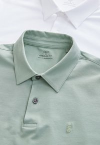 Du mieliniai polo marškiniai, vieni šviesiai žali, o kiti balti, su švelnia tekstūra, mygtukais ir išsiuvinėtu elnio logotipu ant žaliųjų marškinėlių.