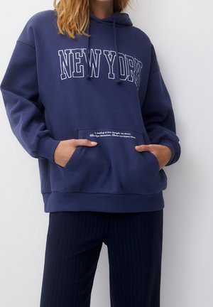 Osoba nosząca granatową bluzę z kapturem z napisem "NEW YORK" z przodu i rękoma w kieszeni typu kangurka, w połączeniu z ciemnymi spodniami w paski.