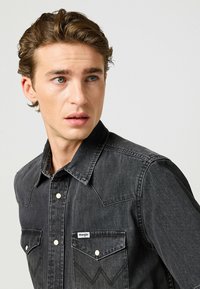Camicia in denim nera con colletto a punta, bottoni automatici e due tasche sul petto. Cuciture sottili e una piccola etichetta "Wrangler" sulla tasca sinistra.