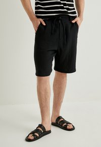 Sorte teksturerede shorts med snøre i taljen og sidelommer, parret med sorte sandaler med remme. Skjorten har vandrette hvide striber.