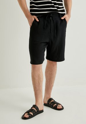 Sorte teksturerede shorts med snøre i taljen og sidelommer, parret med sorte sandaler med remme. Skjorten har vandrette hvide striber.