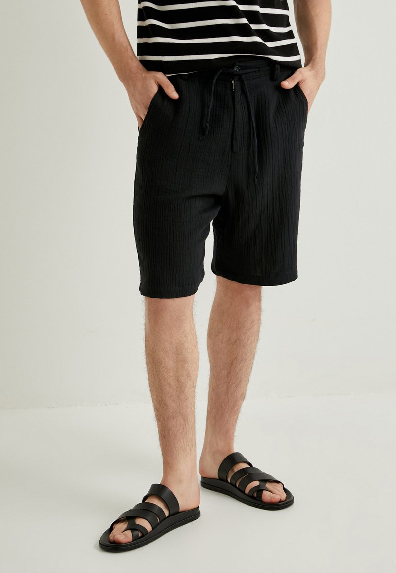 Sorte teksturerede shorts med snøre i taljen og sidelommer, parret med sorte sandaler med remme. Skjorten har vandrette hvide striber.
