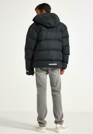 Doudoune noire avec capuche, présentant un design matelassé, des accents réfléchissants et une texture lisse. Associée à un jean gris et des baskets.
