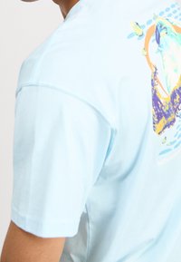 Světle modré bavlněné tričko s krátkými rukávy. Na zadní straně se nachází barevný grafický design s abstraktními tvary a motivem delfína.