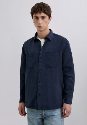 Camisa de botones azul marino hecha de algodón, con cuello, dos bolsillos en el pecho y mangas largas. Combinada con jeans de color azul claro.