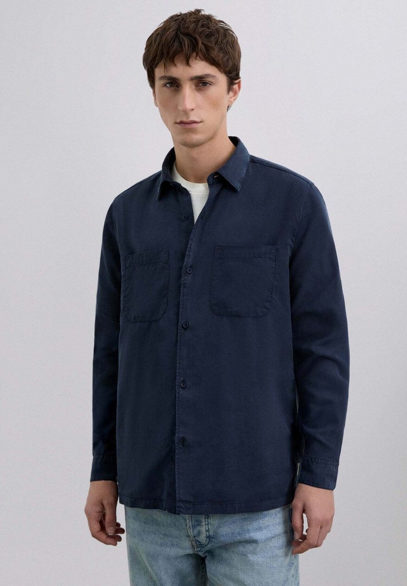 Camisa de botones azul marino hecha de algodón, con cuello, dos bolsillos en el pecho y mangas largas. Combinada con jeans de color azul claro.