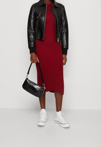 Personne portant une veste en cuir noire, une robe midi rouge, des baskets blanches montantes, et tenant un sac à main noir texturé à côté d'elle.