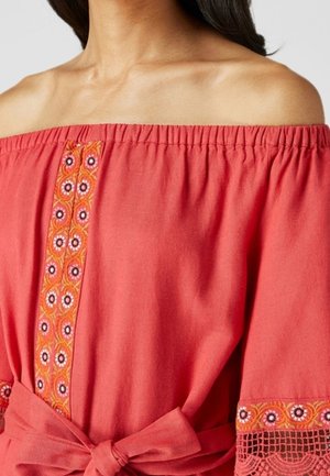 Mujer con una blusa coral estilo off-shoulder con bordado floral en la parte delantera y en los puños de las mangas, atada a la cintura con un cinturón de tela.