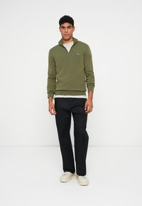 Pullover verde oliva con colletto a zip, abbinato a pantaloni neri e sneakers crema. Tessuto liscio; presenta un piccolo logo sul petto.