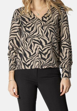 Vrouw draagt een beige en zwarte blouse met zebraprint en lange mouwen en V-hals, met zwarte broek, getoond van nek tot heupen.