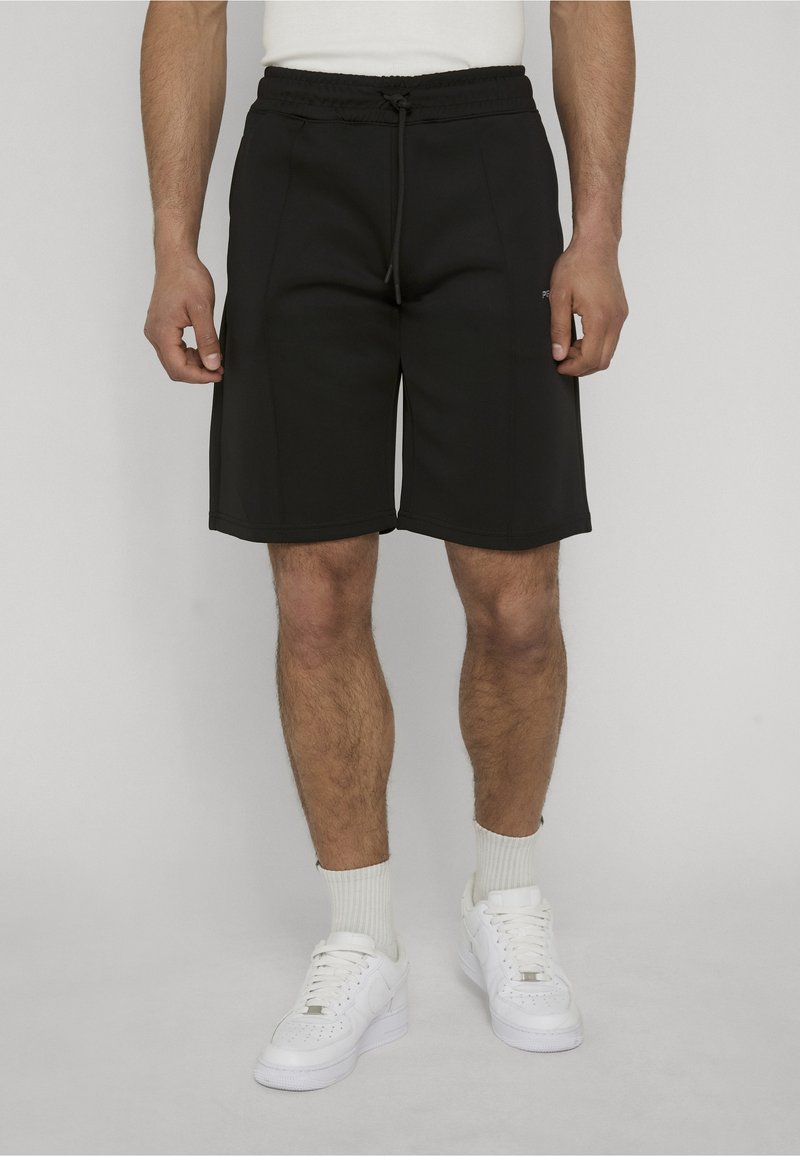 Homme portant un short noir à cordon, des baskets blanches et des chaussettes blanches, debout devant un fond uni.
