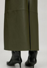 Olijfgroene leren rok met een zijspleet, gecombineerd met zwarte, puntige, hoge hakken, die een gladde textuur en een getailleerd ontwerp benadrukken.