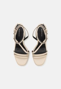 Μπεζ ψηλοτάκουνες σανδάλια με λουράκια και λεία υφή, λεπτά λουράκια στον αστράγαλο και μαύρο εσωτερικό. Διαθέτει τετράγωνη μύτη και μινιμαλιστικό σχέδιο.