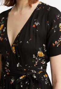 Černé zavinovací šaty s krátkými rukávy, V-výstřihem a květinovým vzorem v žluté, bílé a červené barvě. Texturovaná tkanina s vázaným pasem.