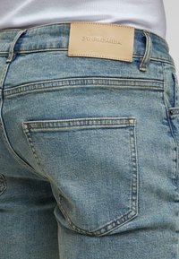 Uvećan prikaz stražnjeg džepa svijetloplavih traperica s bež kožnom oznakom brenda i bijelom košuljom ugrnutom unutra.