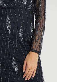 Námořnické šaty s dlouhými průsvitnými rukávy, zdobené stříbrnými flitrovými vlnovkovými vzory a přiléhavým střihem. Texturovaná látka s podrobnými ozdobami.