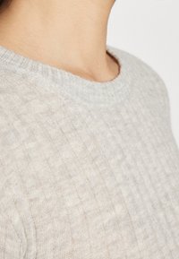 Primo piano di una persona che indossa un maglione a coste beige chiaro con scollatura rotonda su uno sfondo neutro.