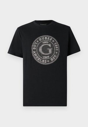 Svart bomulls T-skjorte med rund grafikk i hvitt med bokstaven "G" og teksten "GUESS EST. 1981" i et horisontalt mønster. Korte ermer.