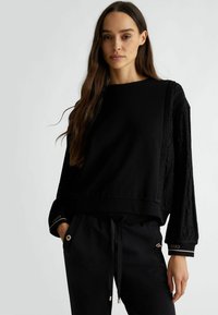 Felpa nera con maniche nere testurizzate, scollatura rotonda e orlo elasticizzato. Presenta dettagli dorati ai polsini, abbinata a pantaloni neri.