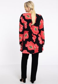 Tunique noire avec un grand motif floral rouge, à manches longues et à coupe ample, associée à un pantalon noir et des chaussures à talons hauts, vue de dos.
