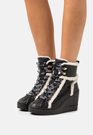 Bottines compensées noires à cheville avec bordure en fausse fourrure blanche, lacets marqués "Guess" et logo latéral embossé, portées sur des jambes nues sur fond blanc.