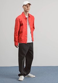 Sarkans kokvilnas žakets ar spiedpogām, baltā t-kreklā apakšā, melnas platās bikses un baltas kedas. Stāv uz zilā paklāja.