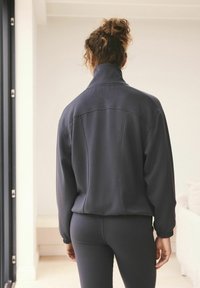 Tmavě šedá zipová bunda s vysokým límcem, vyrobená z texturované látky a s elastickými manžetami, nošená s odpovídajícími legínami, viděná zezadu.