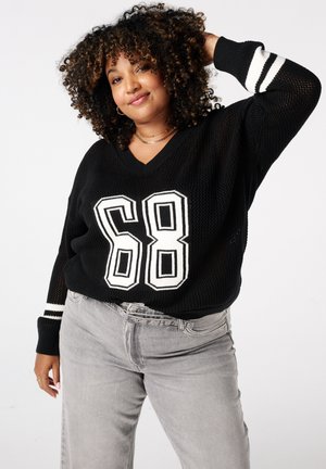 Femme aux cheveux bouclés portant un pull noir avec le numéro "68" en blanc et un pantalon gris, posant une main derrière la tête contre un fond blanc.