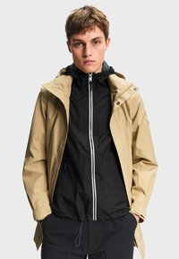 Jeune homme portant une veste de pluie beige par-dessus un sweat à capuche zippé noir, debout les mains dans les poches devant un fond uni.