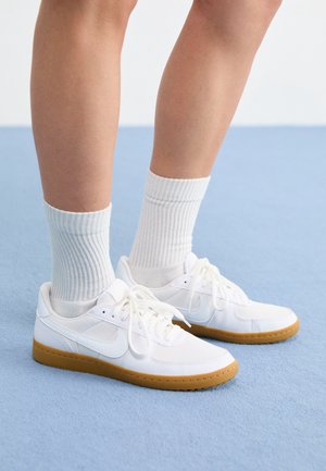 Jambes portant des chaussettes blanches côtelées et des baskets en cuir blanches avec des semelles en gomme, debout sur un sol tapissé bleu clair.